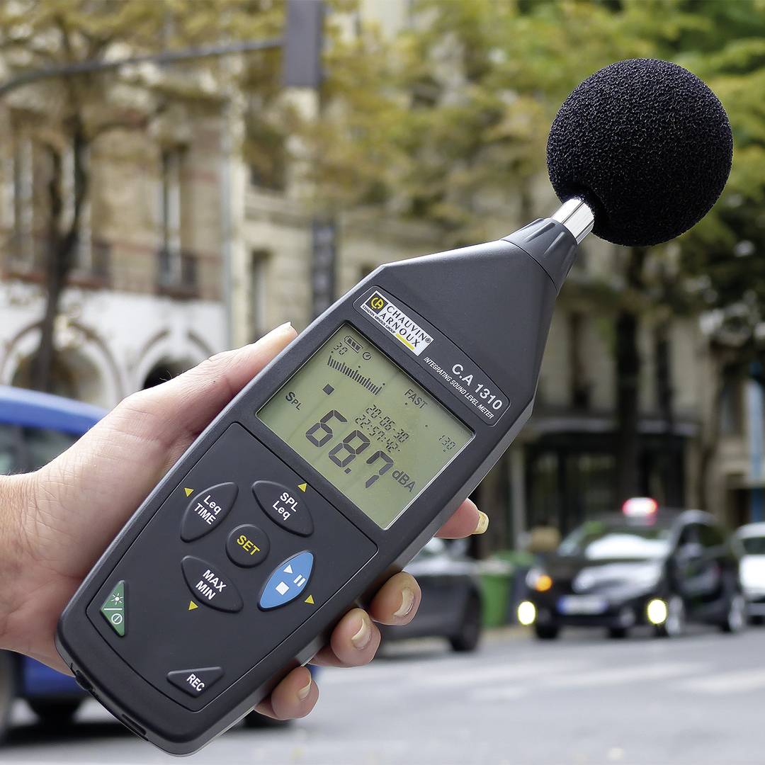 Eine Hand hält ein Schallpegelmessgerät, das 68,7 dB anzeigt, auf einer Stadtstraße mit vorbeifahrenden Autos und Bäumen im Hintergrund.