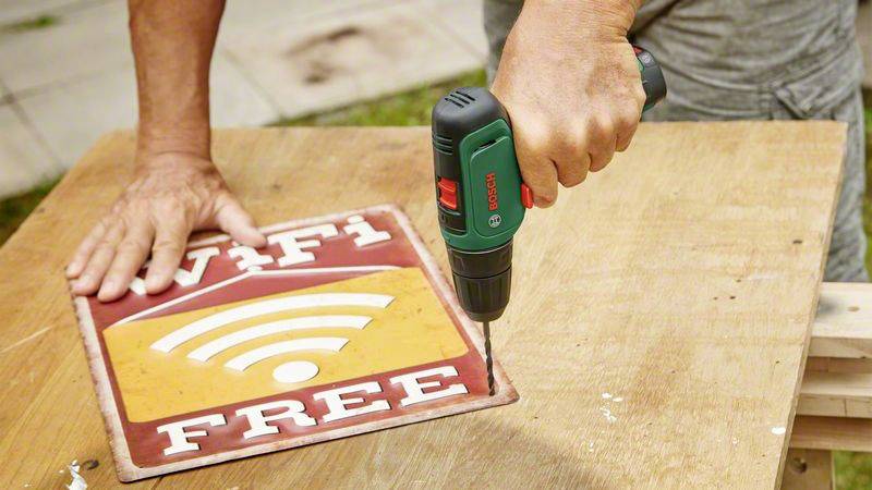 Eine Person befestigt ein Schild mit der Aufschrift 'WiFi Free' mit einem Akkuschrauber auf einem Holztisch.