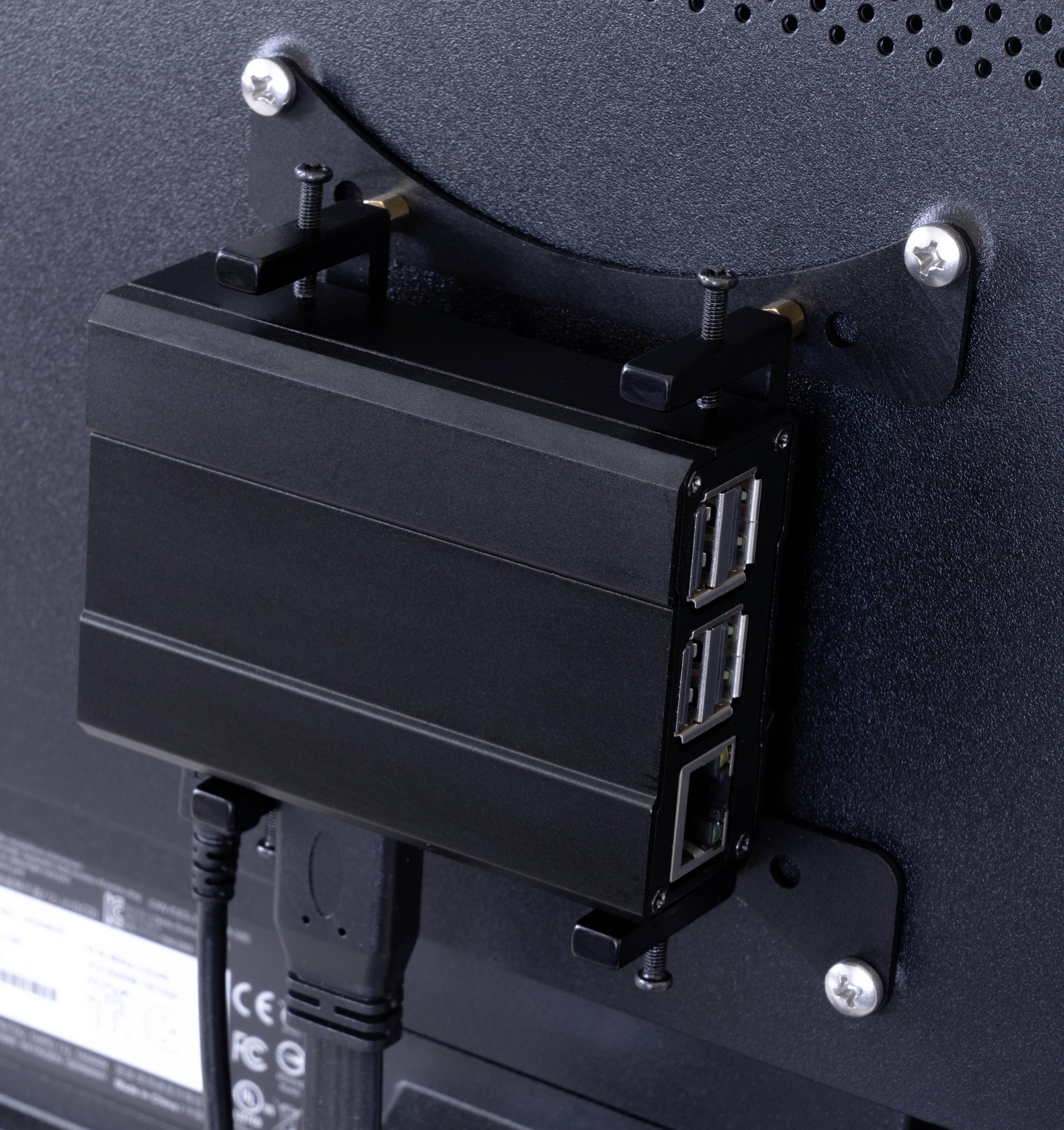 Ein kleiner schwarzer Computer mit mehreren USB- und Ethernet-Anschlüssen ist an der Rückseite eines Monitors montiert.