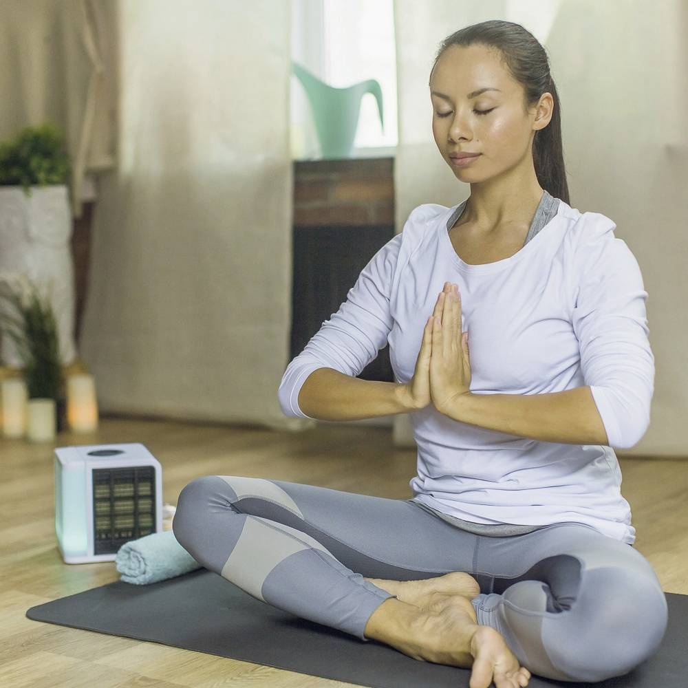 Eine Frau in Meditationspose sitzt auf einer Yogamatte in einem Raum mit Kerzen und Pflanzen, die Ruhe und Entspannung ausstrahlt.