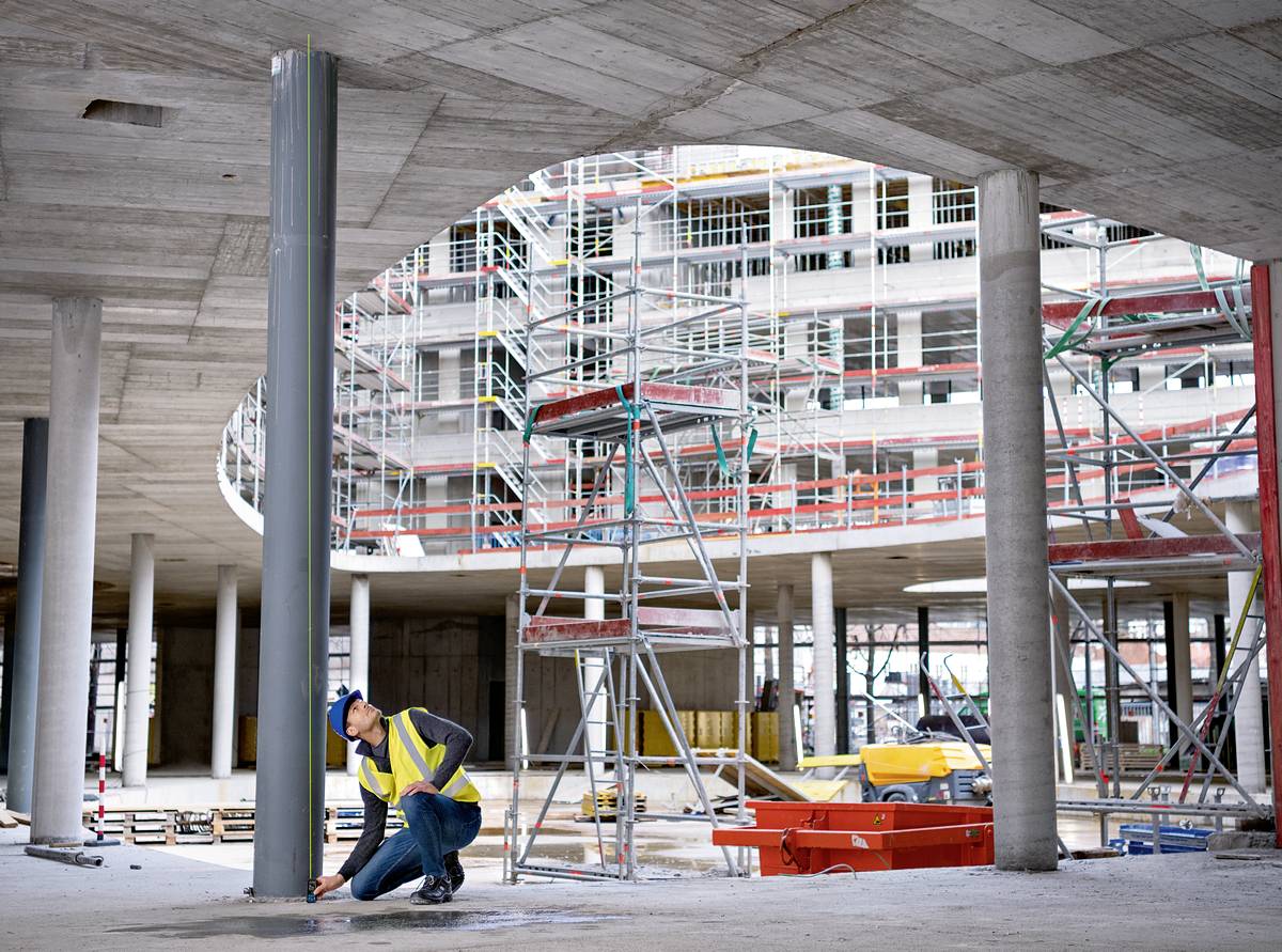Eine Bauarbeiterin in gelber Warnweste überprüft einen Betonpfeiler auf einer Baustelle mit Gerüsten und unfertigem Gebäude im Hintergrund.