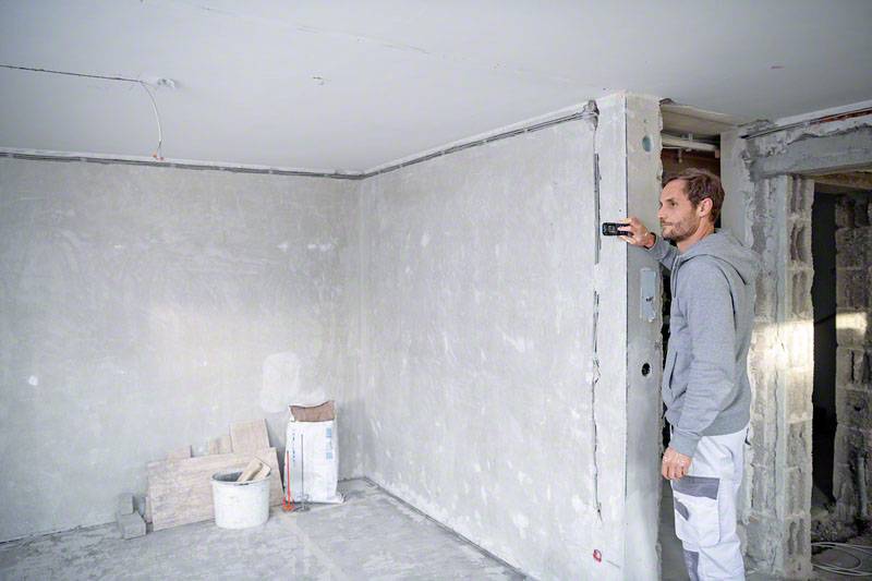 Ein Mann in Arbeitskleidung misst eine Wand in einem unfertigen Raum mit einem Laserentfernungsmesser. Baustellenmaterial ist im Hintergrund.
