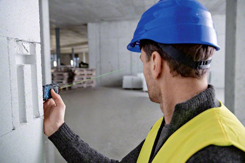 Ein Bauarbeiter mit blauem Helm und gelber Weste verwendet ein Laser-Entfernungsmessgerät an einer Betonwand in einer Baustelle.