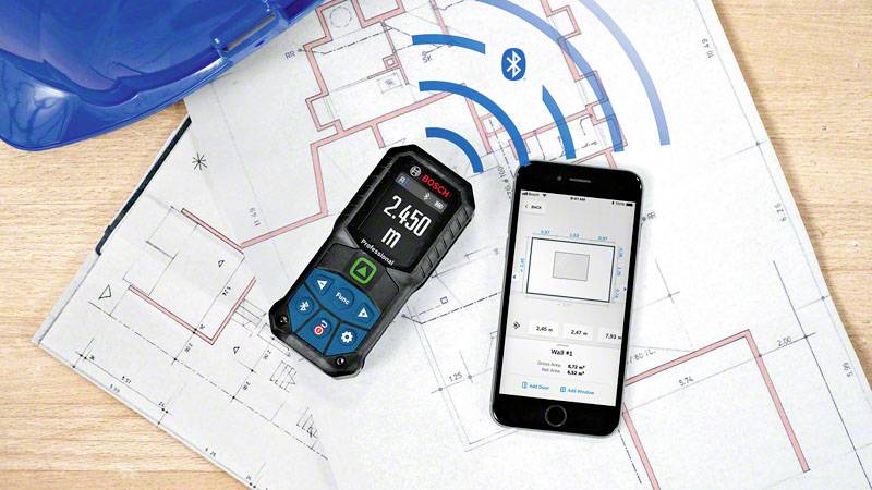 Smartphone und Laserentfernungsmesser auf Bauplänen, verbunden via Bluetooth. Der Laser zeigt '2,450 m' an, App zeigt Messung.