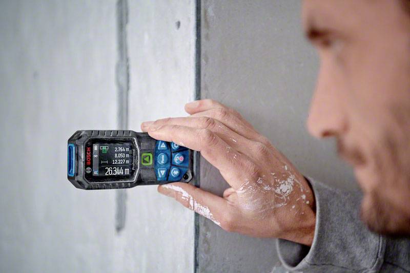 Eine Person misst mit einem digitalen Laser-Entfernungsmesser die Länge einer Betonwand. Das Gerät zeigt '2634 m' an.