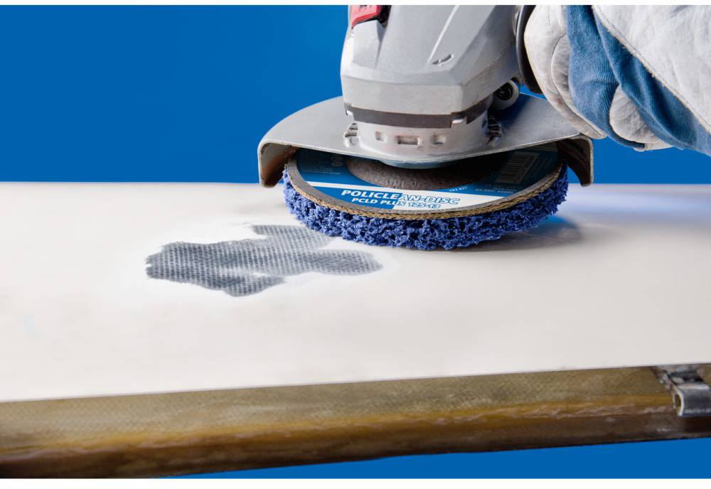 Ein Schleifgerät glättet die Oberfläche eines weißen Brettes. Ein Arbeiter hält das Gerät, das Staubpartikel erzeugt, auf die Fläche.