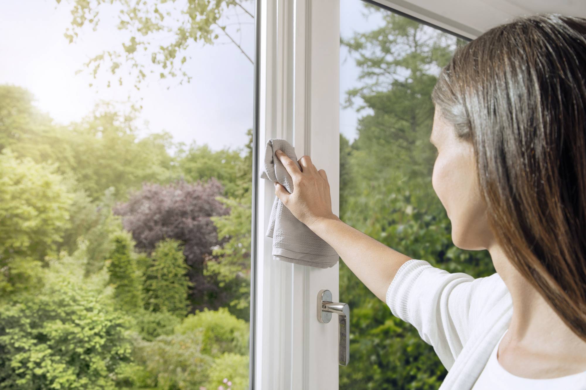 Frau reinigt von innen ein großes Fenster mit Aussicht auf einen sonnigen, grünen Garten. Sie verwendet ein graues Tuch.