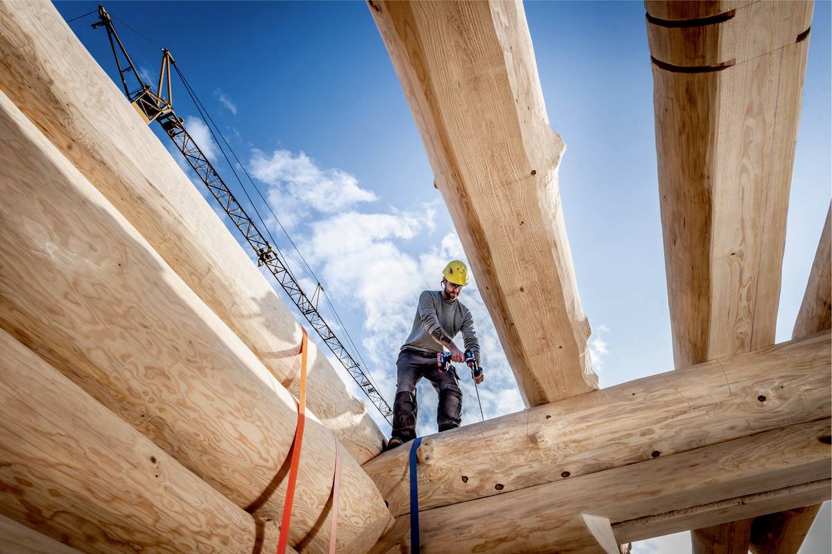 Ein Arbeiter mit Schutzhelm steht auf einem Holzbalken und nutzt ein Werkzeug. Im Hintergrund ein Baukran und blauer Himmel.