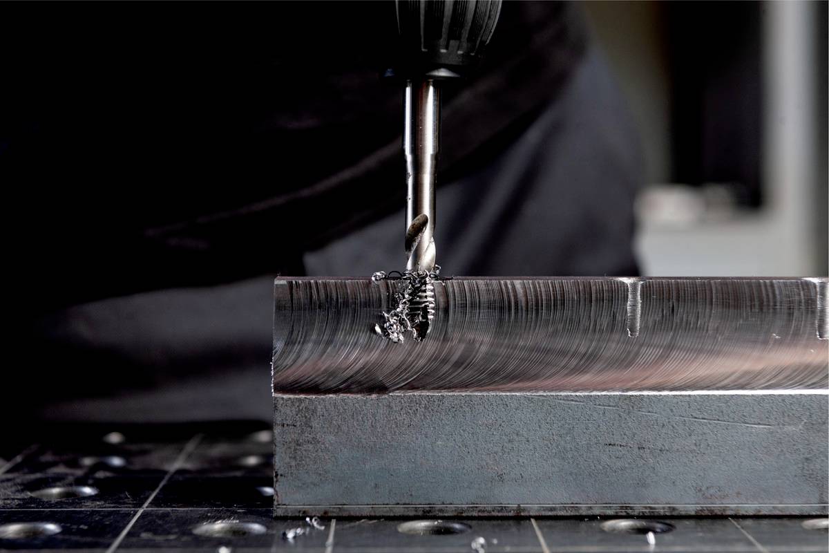 Ein Metallbohrer bearbeitet ein Stück Metall auf einem Tisch, dabei entstehen Späne. Die Szene zeigt Präzisionsarbeit in einem Werkstattkontext.