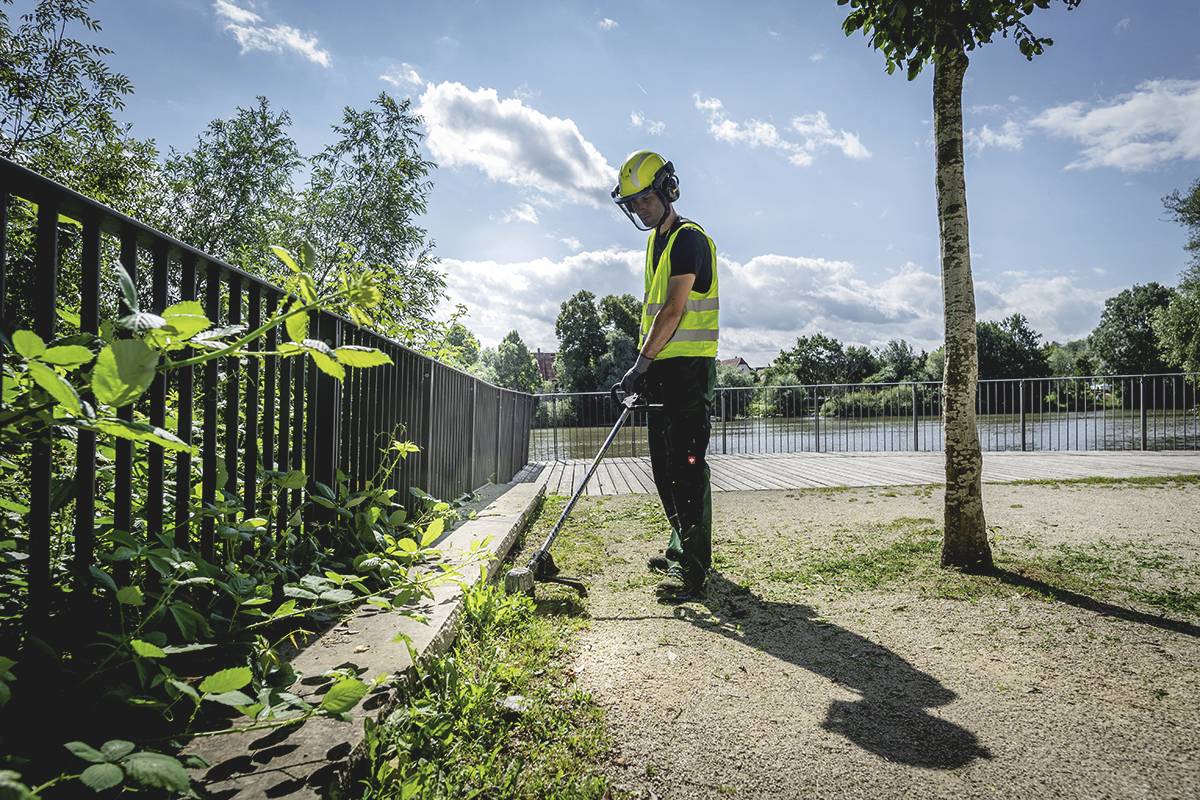 Eine Person in Sicherheitskleidung verwendet einen Rasentrimmer, um Pflanzen an einem Zaun in einem Park nahe an einem Fluss zu trimmen.