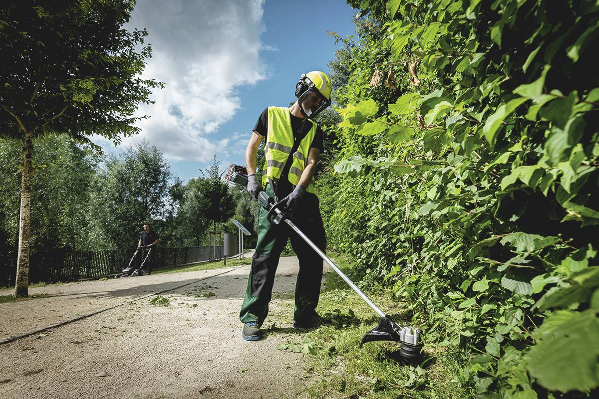 Ein Arbeiter mit Schutzkleidung schneidet Büsche mit einem Trimmer neben einem Gehweg. Ein weiterer Arbeiter im Hintergrund.