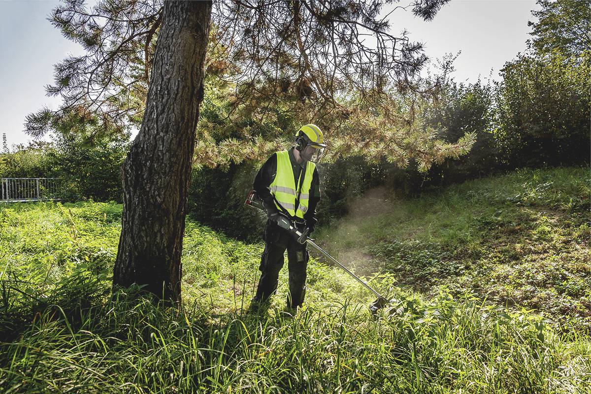 Eine Person mäht hohes Gras unter einem Baum mit einem Rasentrimmer. Sie trägt Schutzkleidung mit Warnweste und Helm.