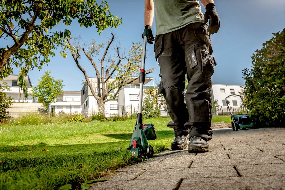 Eine Person mäht mit einem Rasenkantenschneider den Rasen entlang eines gepflasterten Weges in einem sonnigen Garten.