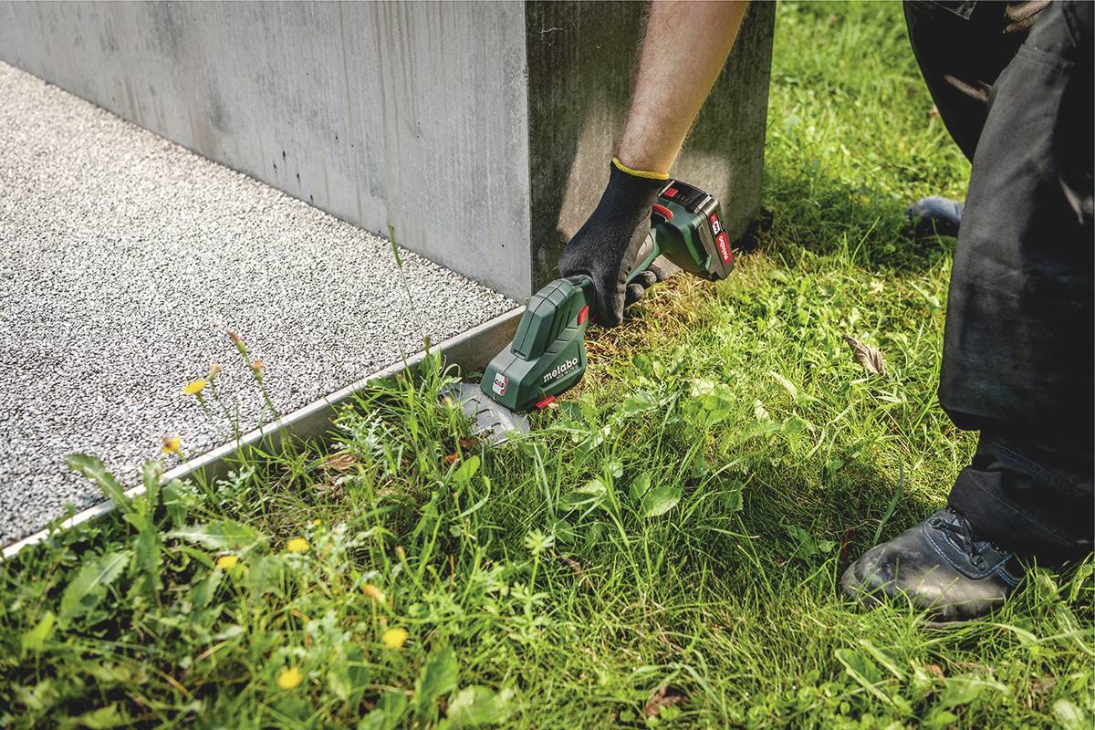 Person schneidet mit einer elektrischen Gras- und Strauchschere Rasen an einer Kante ab. Florierende Gräser und Blumen sind um das Gerät sichtbar.