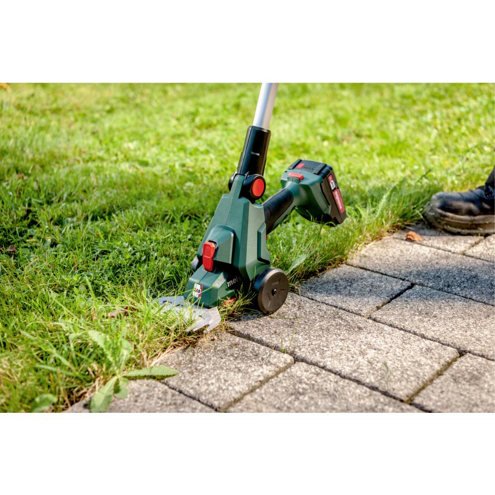 Ein schnurloser Rasentrimmer wird verwendet, um Gras entlang eines Steinweges zu schneiden. Der Trimmer hat einen grünen Körper und scheint batteriebetrieben zu sein.