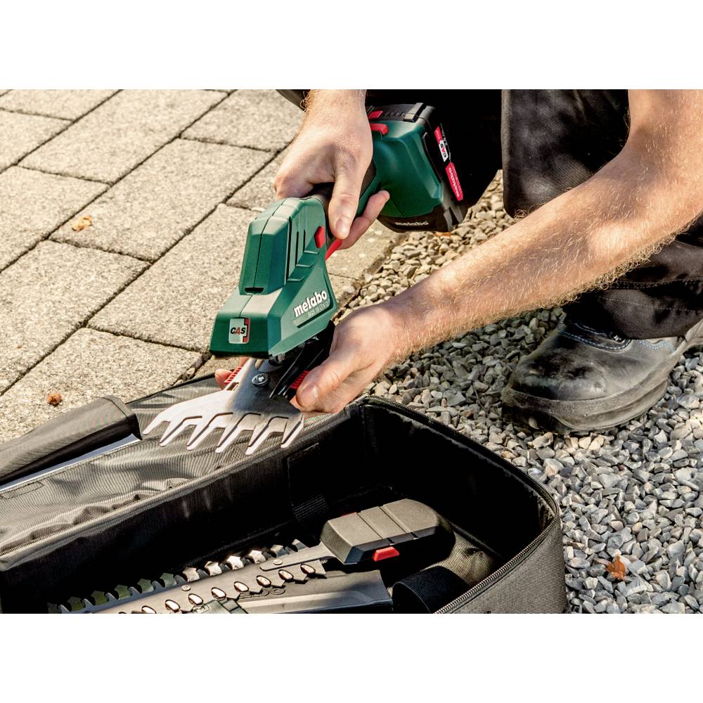 Eine Person justiert die Klinge eines grünen Elektrowerkzeugs, wobei ein Kies- und Pflasterbelag im Hintergrund zu sehen ist. Eine Tasche mit zusätzlichen Werkzeugen befindet sich in der Nähe.