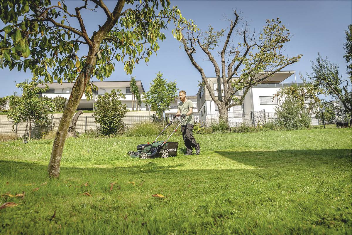 Ein Mann mäht Rasen in einem sonnigen Garten. Im Hintergrund sind moderne Häuser und Bäume zu sehen.