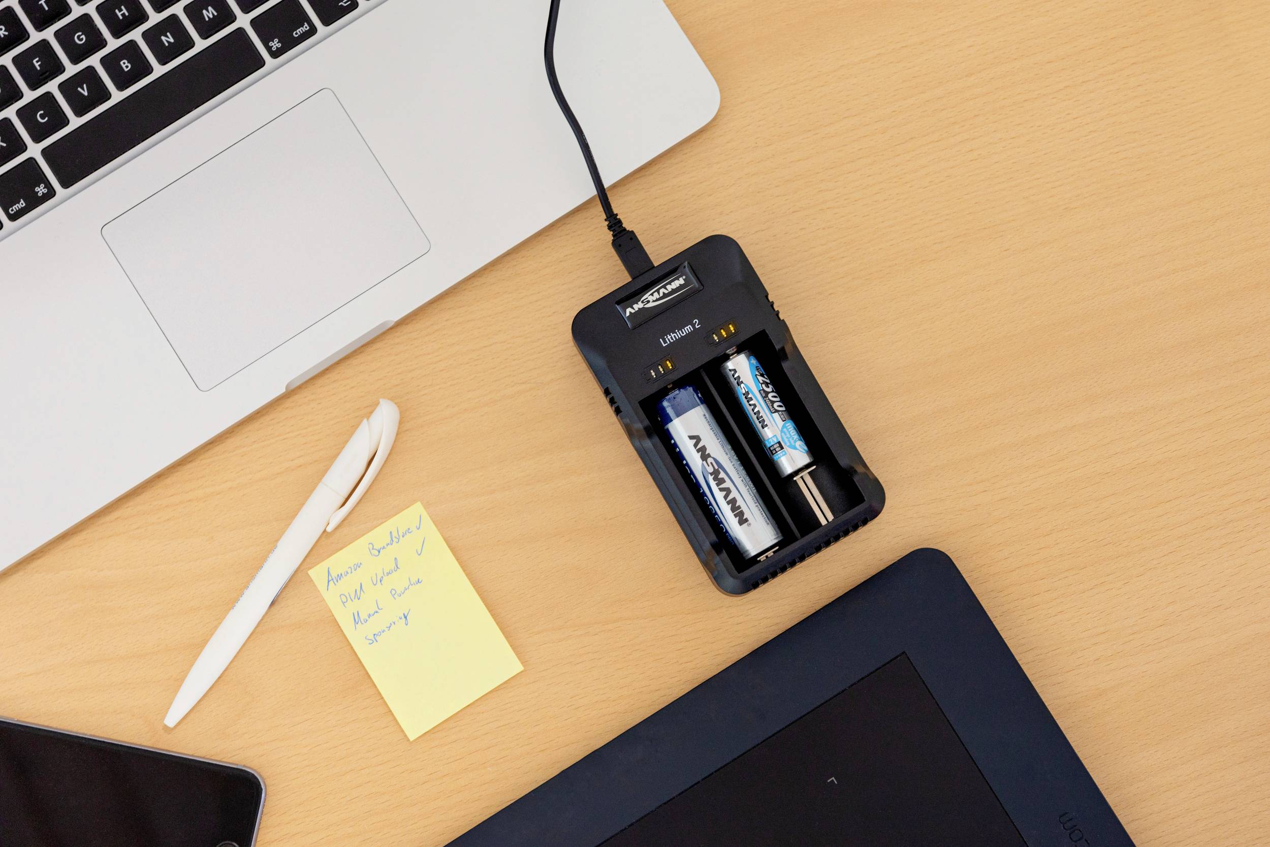 Ein Tisch mit einem Laptop, einem Stift, einem gelben Notizzettel neben einem Akkuladegerät mit zwei verschiedenen AA-Batterien.