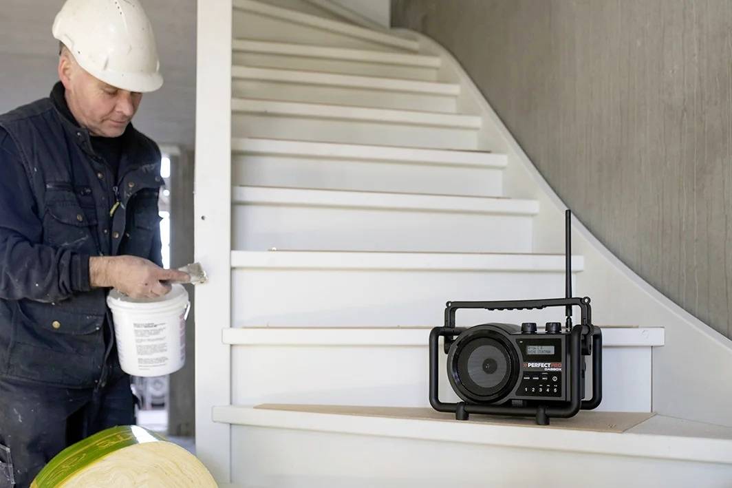 Ein Bauarbeiter mit weißem Helm streicht eine Treppe. Auf der Treppe steht ein Baustellenradio.