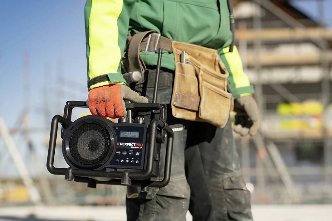 Ein Bauarbeiter in Arbeitskleidung hält ein tragbares Radio auf einer Baustelle. Im Hintergrund sind unscharf Baugerüste zu sehen.