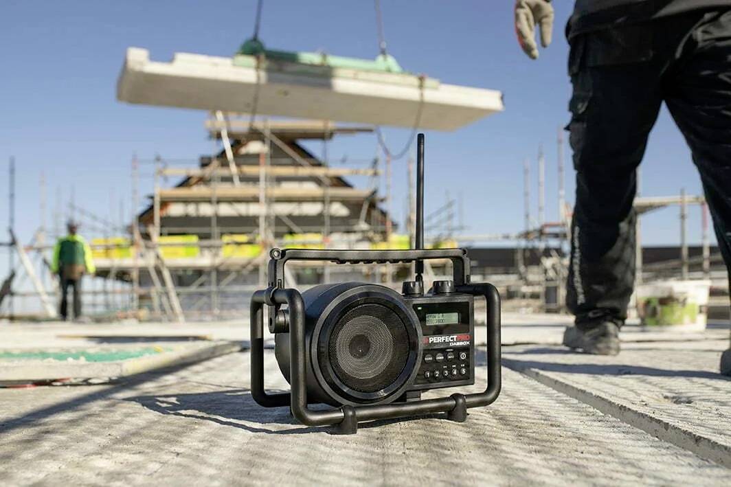 Baustelle mit portablem Radio im Vordergrund, Arbeiter und Baugerüst im Hintergrund; Gebäude im Bau; sonniger Tag.