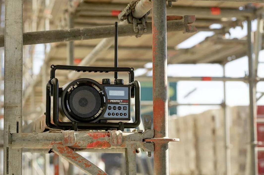 Ein tragbares Radio steht auf einem Baugerüst. Es ist robust gebaut, mit einer Antenne und mehreren Bedientasten, in einer Baustellenumgebung.