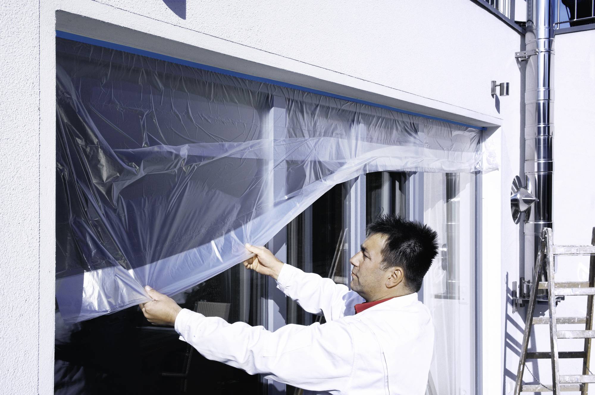 Ein Mann in Arbeitskleidung befestigt eine Plastikfolie an einem großen Fenster eines weißen Gebäudes. Außenbereich mit Tageslicht.