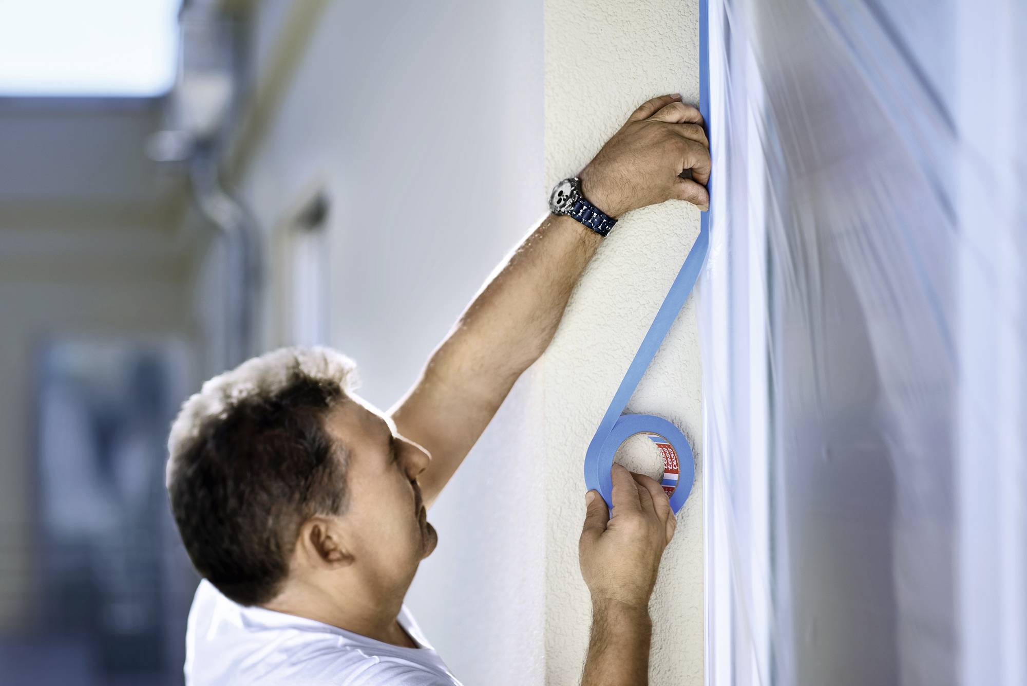 Eine Person befestigt blaues Klebeband an einer Außenwand. Sie trägt ein weißes T-Shirt und eine Uhr. Sie konzentriert sich auf die Arbeit.