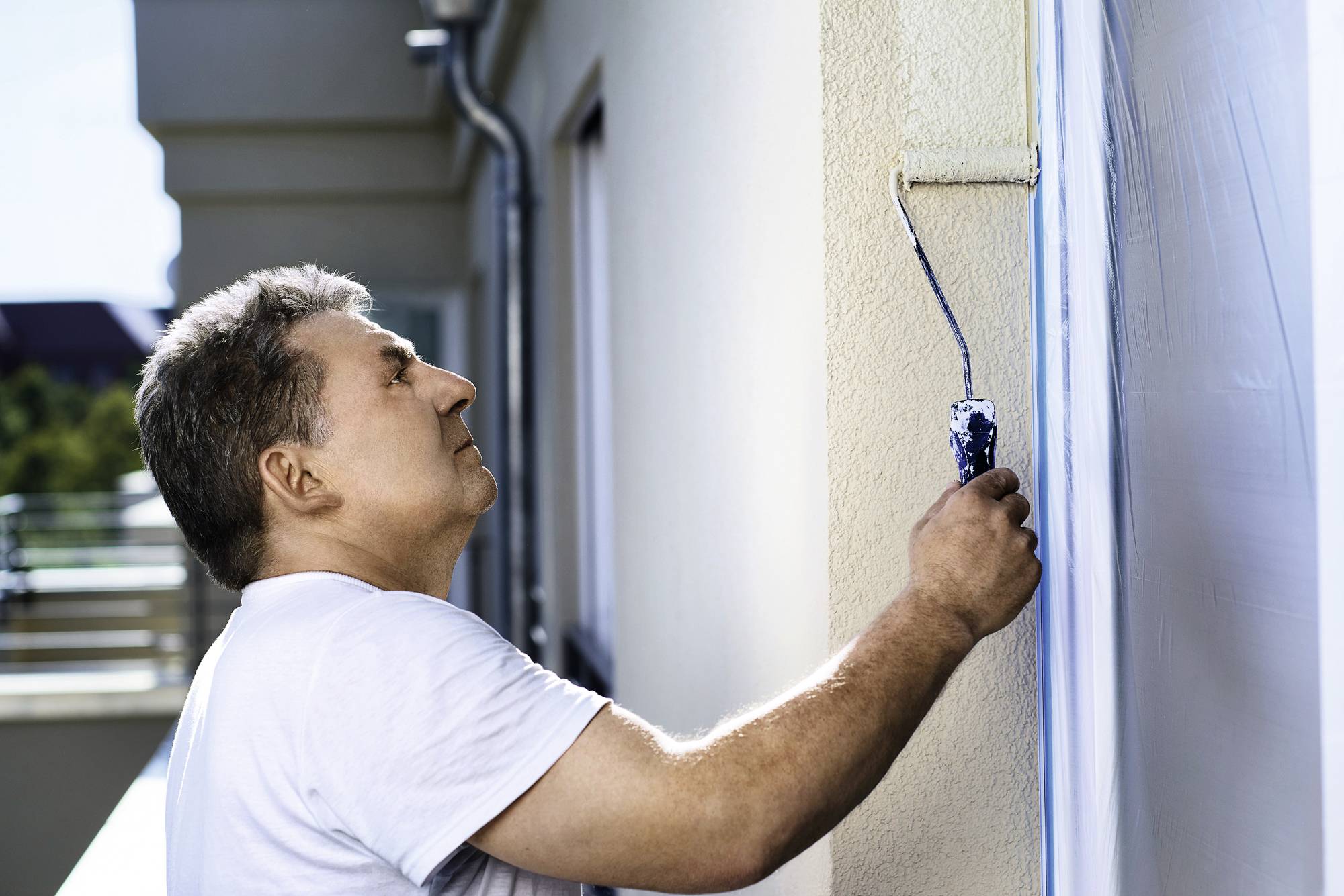Ein Mann streicht mit einer Farbrolle eine Hauswand. Er konzentriert sich auf seine Arbeit und trägt ein weißes T-Shirt.
