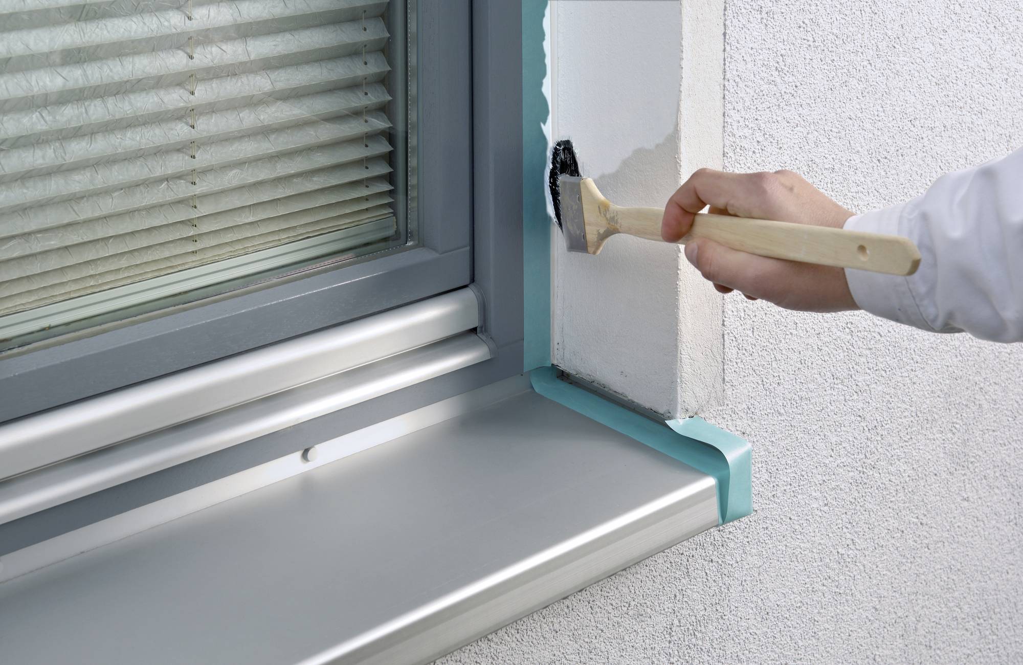 Eine Hand streicht weiße Farbe auf die Wand um einen Fensterrahmen. Das Fenster hat Jalousien, und der Rahmen ist teilweise mit Klebeband geschützt.