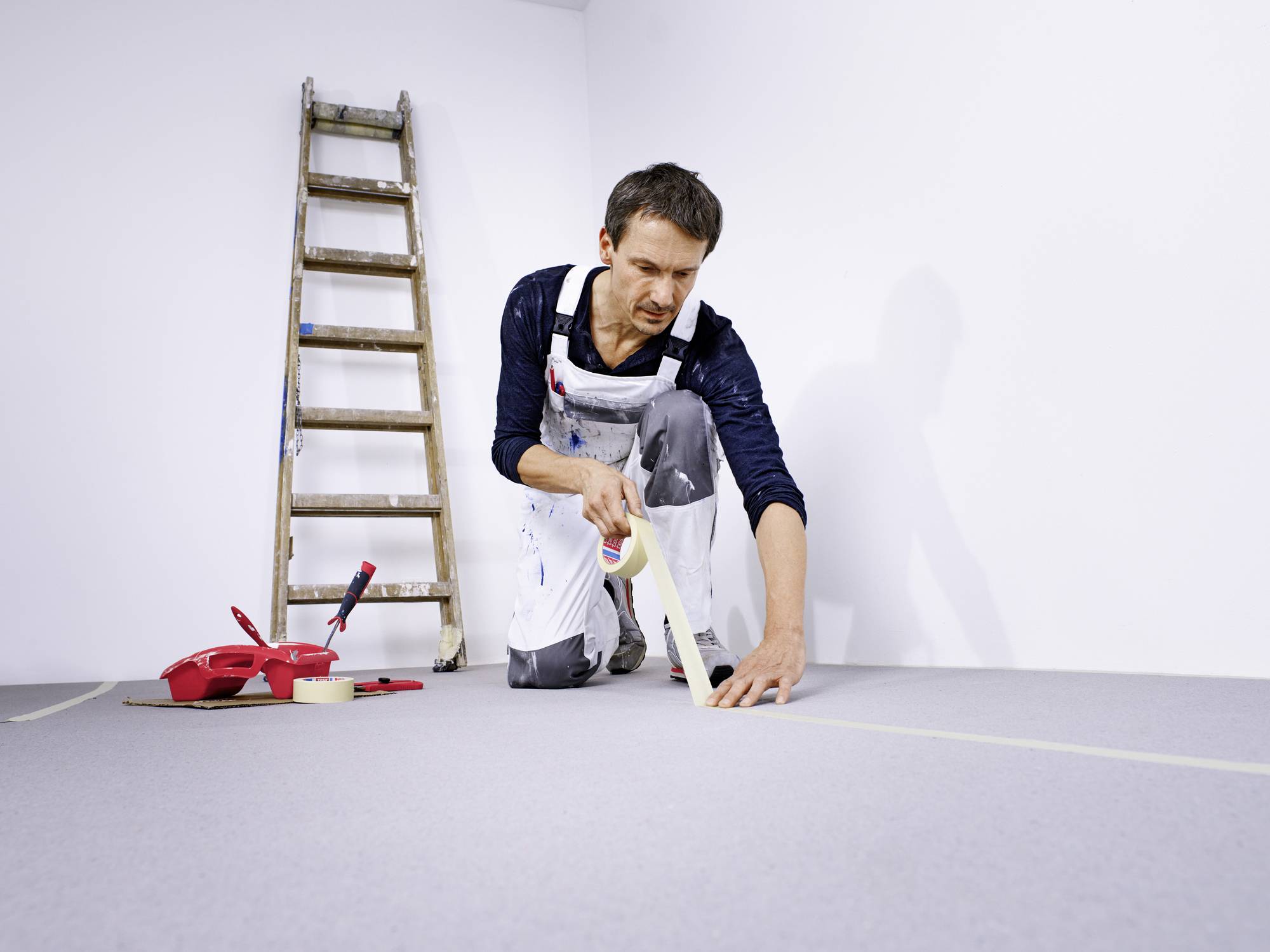 Ein Maler in Arbeitskleidung klebt den Boden mit Malerband ab. Im Hintergrund steht eine Leiter.