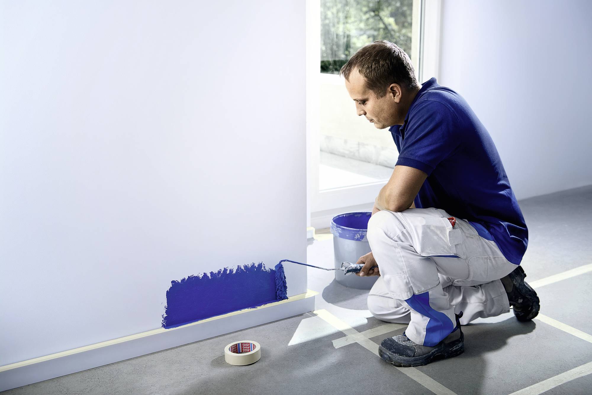 Ein Mann in Arbeitskleidung streicht eine Wand mit blauer Farbe. Neben ihm steht ein Farbeimer, und Malerkrepp klebt auf dem Boden.
