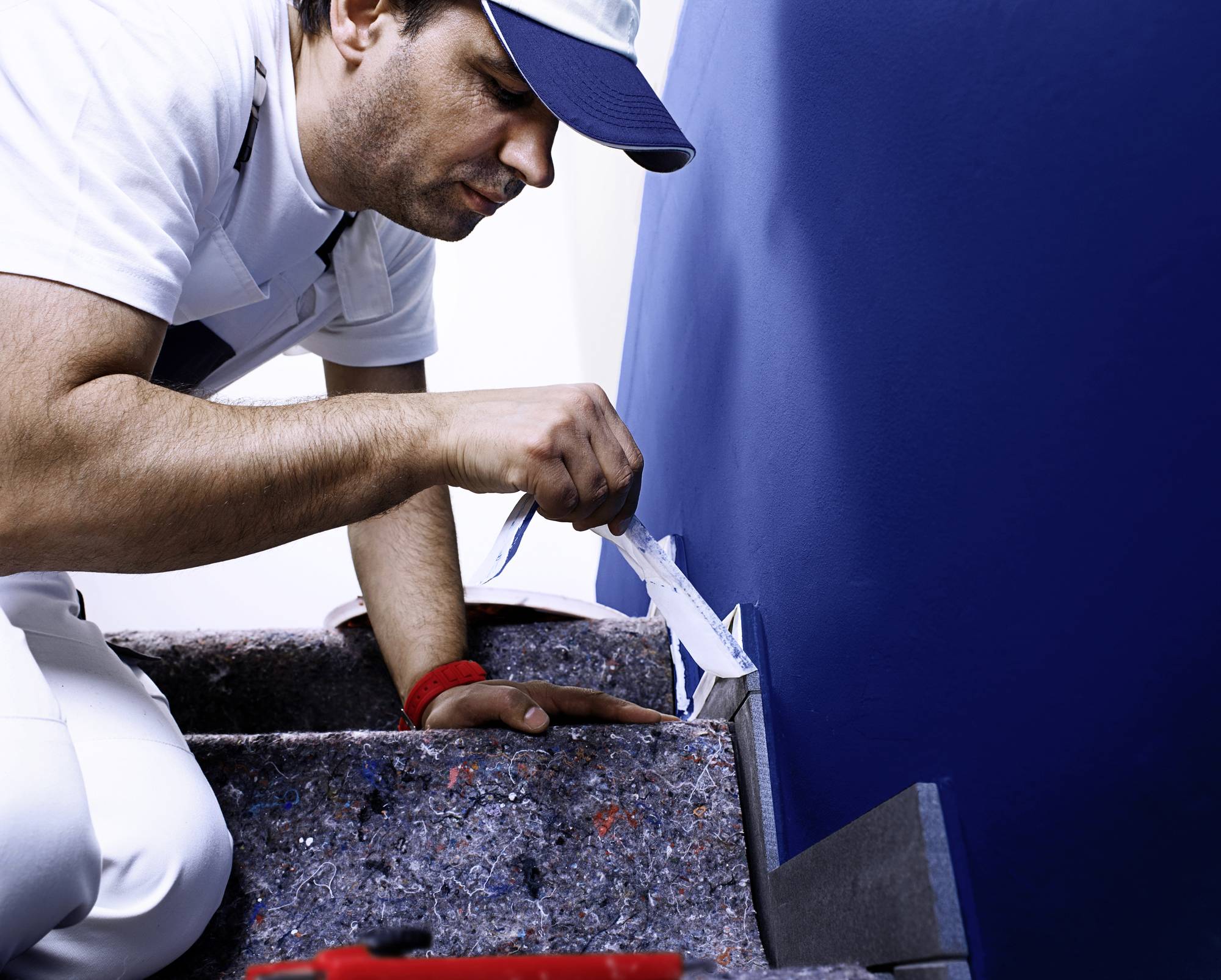 Ein Maler in Arbeitskleidung trägt blaue Farbe auf eine Wand auf. Er benutzt einen Pinsel und arbeitet an Kanten sorgfältig.