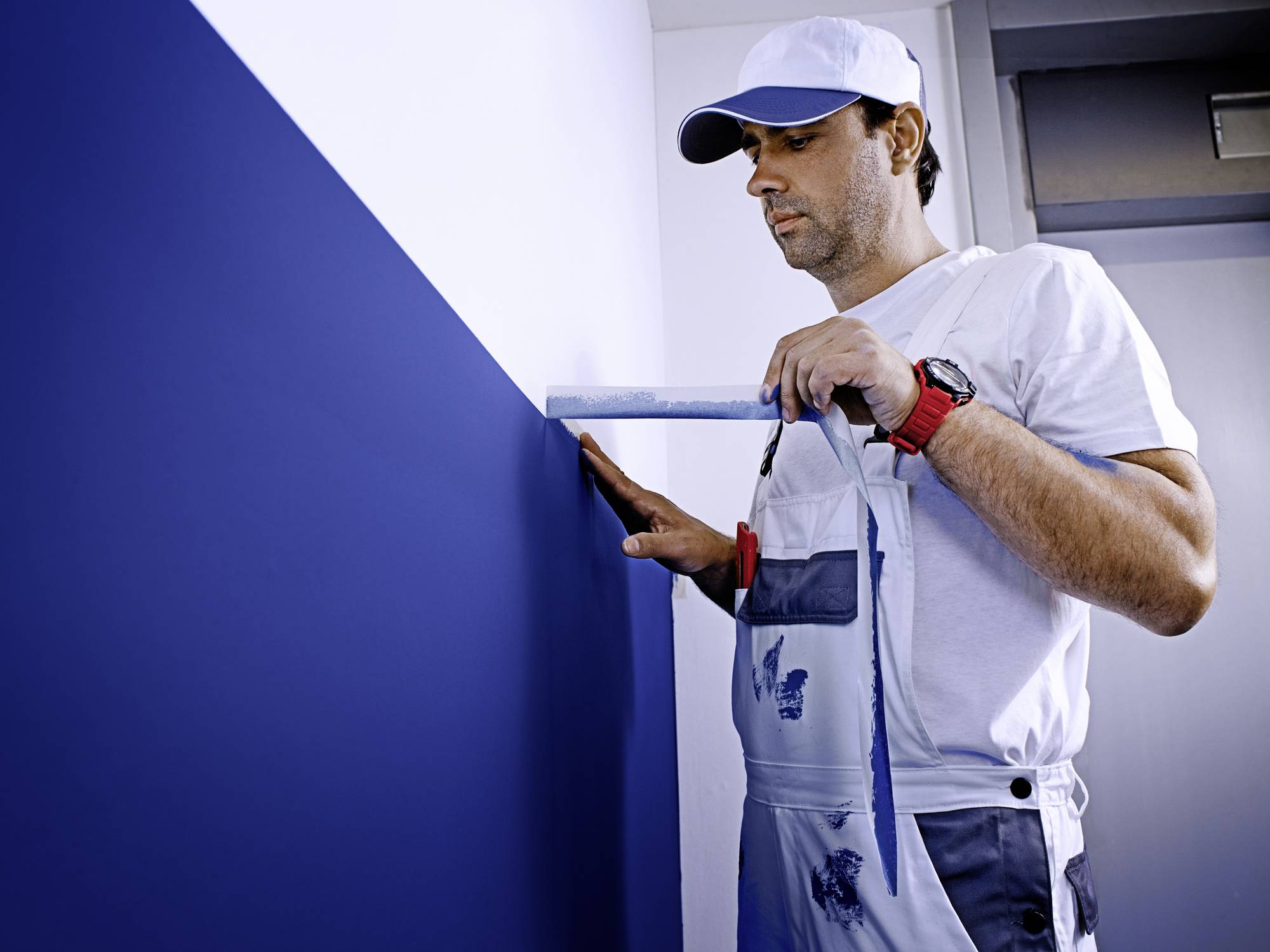 Ein Mann im Overall misst mit einem Lineal die Höhe der blauen Wand. Er trägt eine weiße Kappe und eine Uhr.