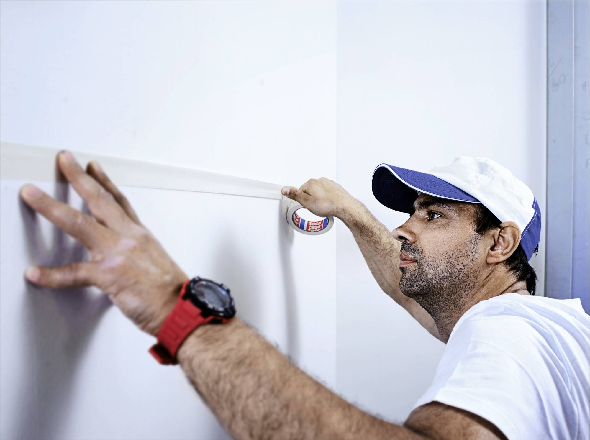 Ein Mann klebt vorsichtig Malerband an eine weiße Wand. Er trägt ein weißes T-Shirt und eine Kappe. Er arbeitet konzentriert.