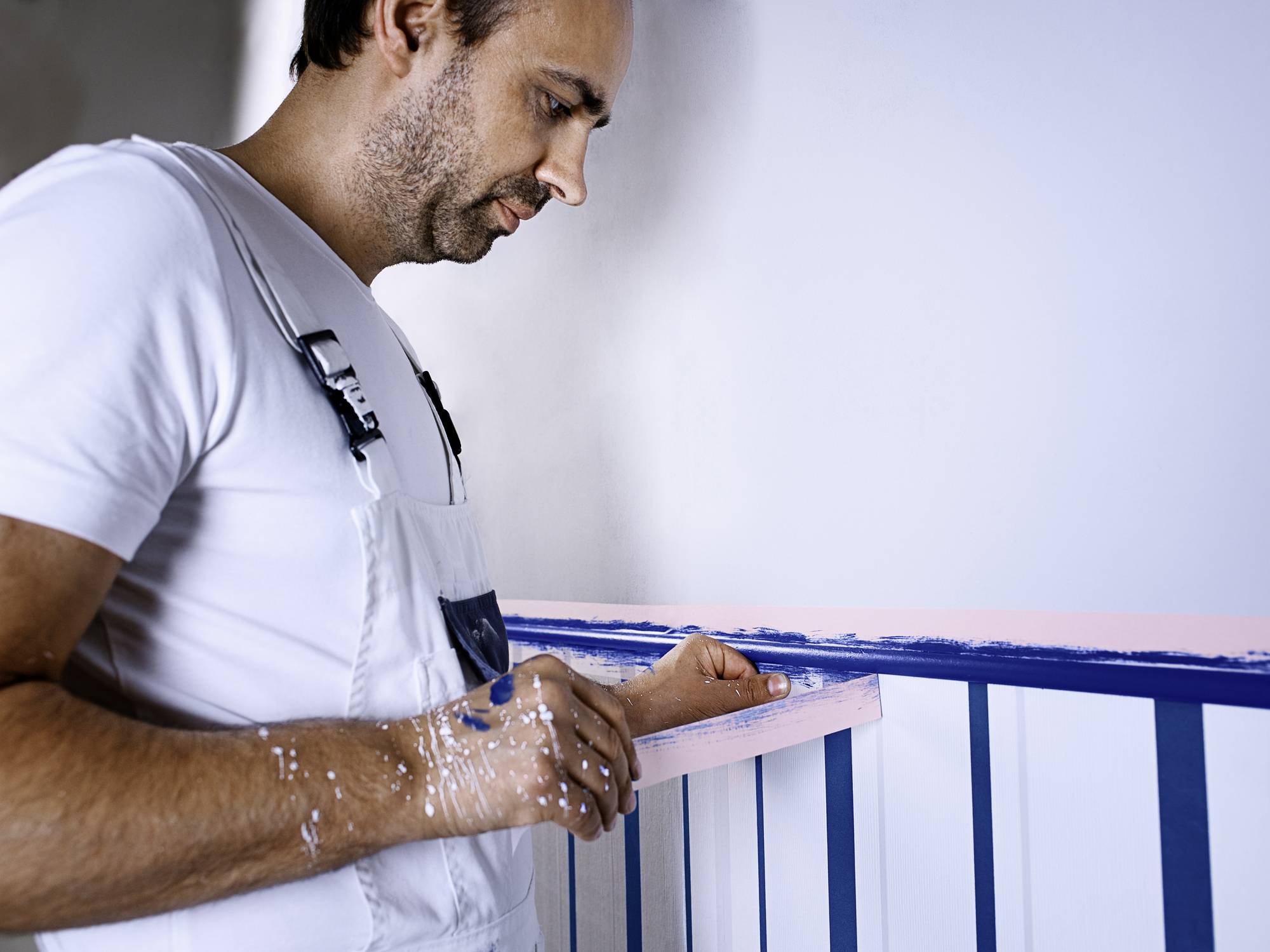 Ein Mann in Arbeitskleidung streicht sorgfältig ein Balkongeländer mit blauer Farbe. Sein Pinsel und Hände sind mit Farbe bedeckt.