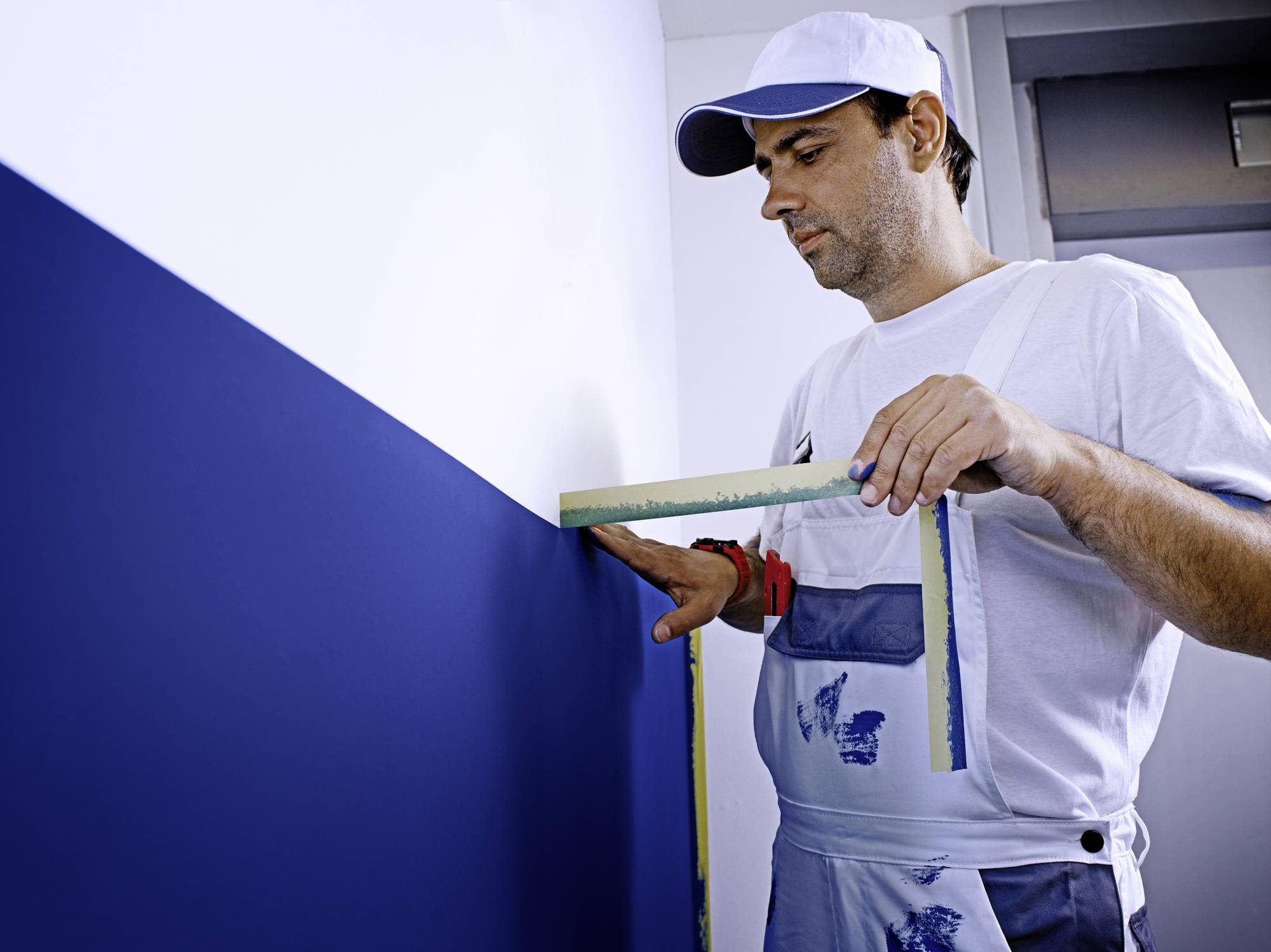 Ein Mann in Arbeitskleidung misst mit einem Maßband eine blau gestrichene Wand. Er trägt eine weiße Mütze und eine Hose mit Farbflecken.