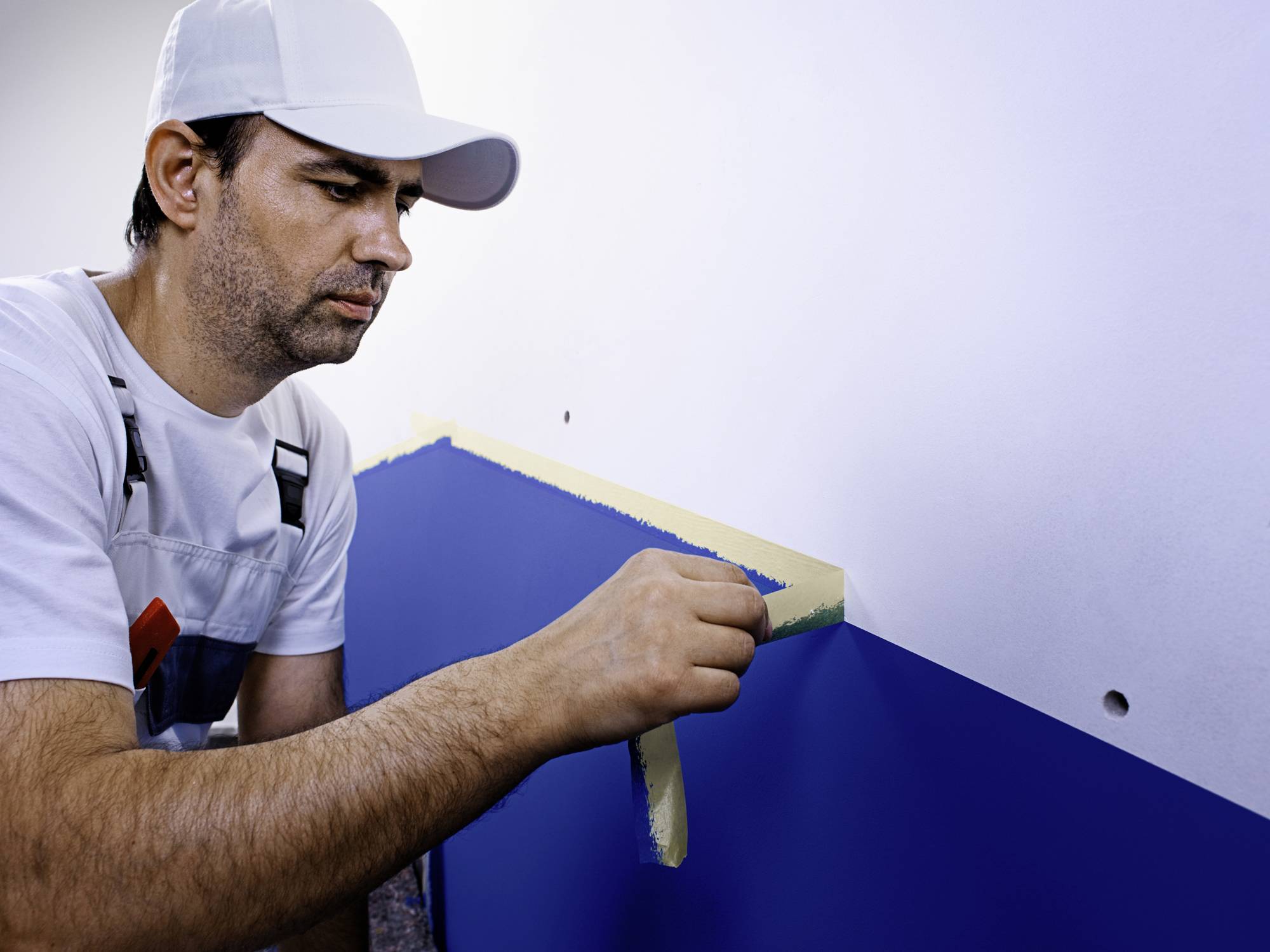 Ein Maler in Arbeitskleidung klebt mit einem Klebeband eine Wand ab, um diese zweifarbig zu streichen. Die Wand ist teilweise blau.