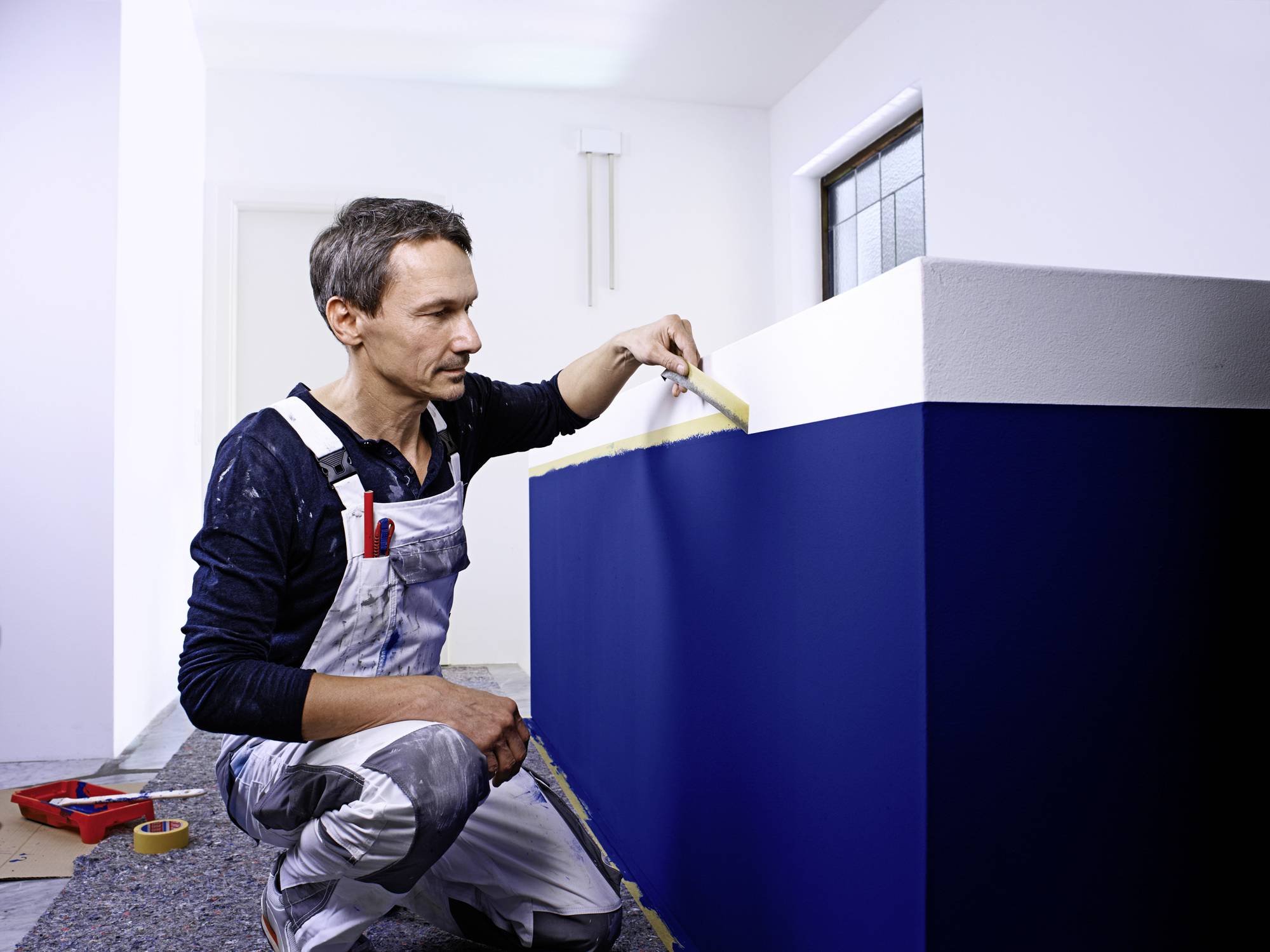 Maler in Arbeitskleidung streicht eine blaue Wand mit einem Pinsel. Im Hintergrund ist ein weißer Raum zu sehen.
