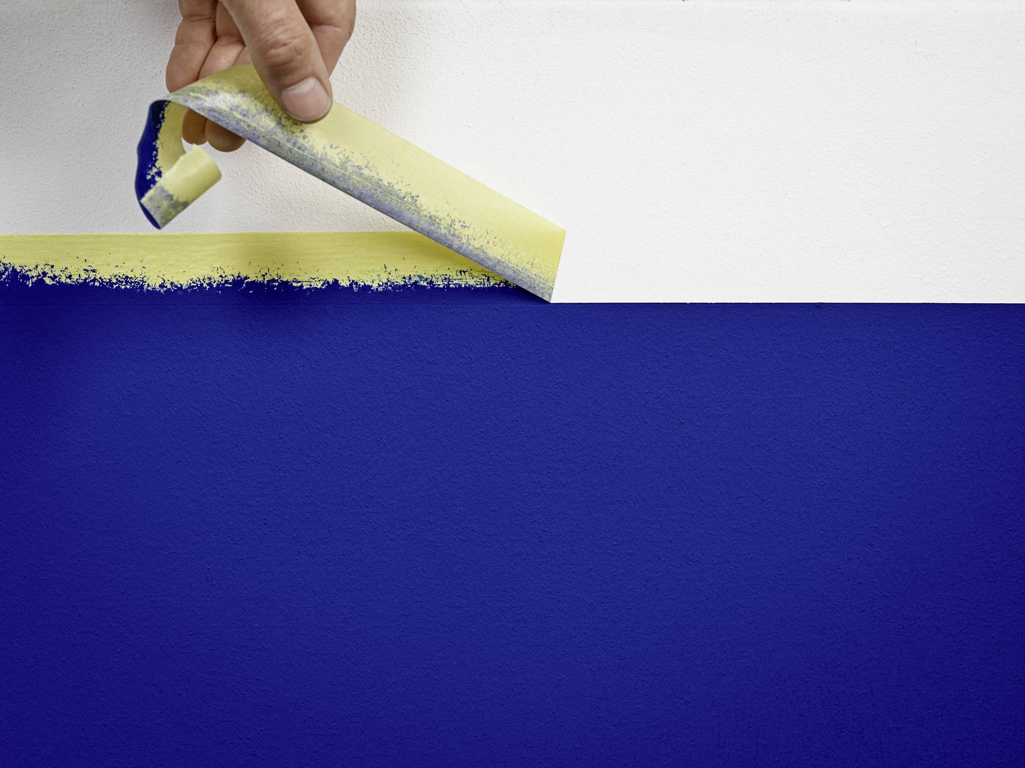 Eine Hand entfernt Klebeband von einer Wand und enthüllt eine klare Grenze zwischen weißer und blauer Farbe.