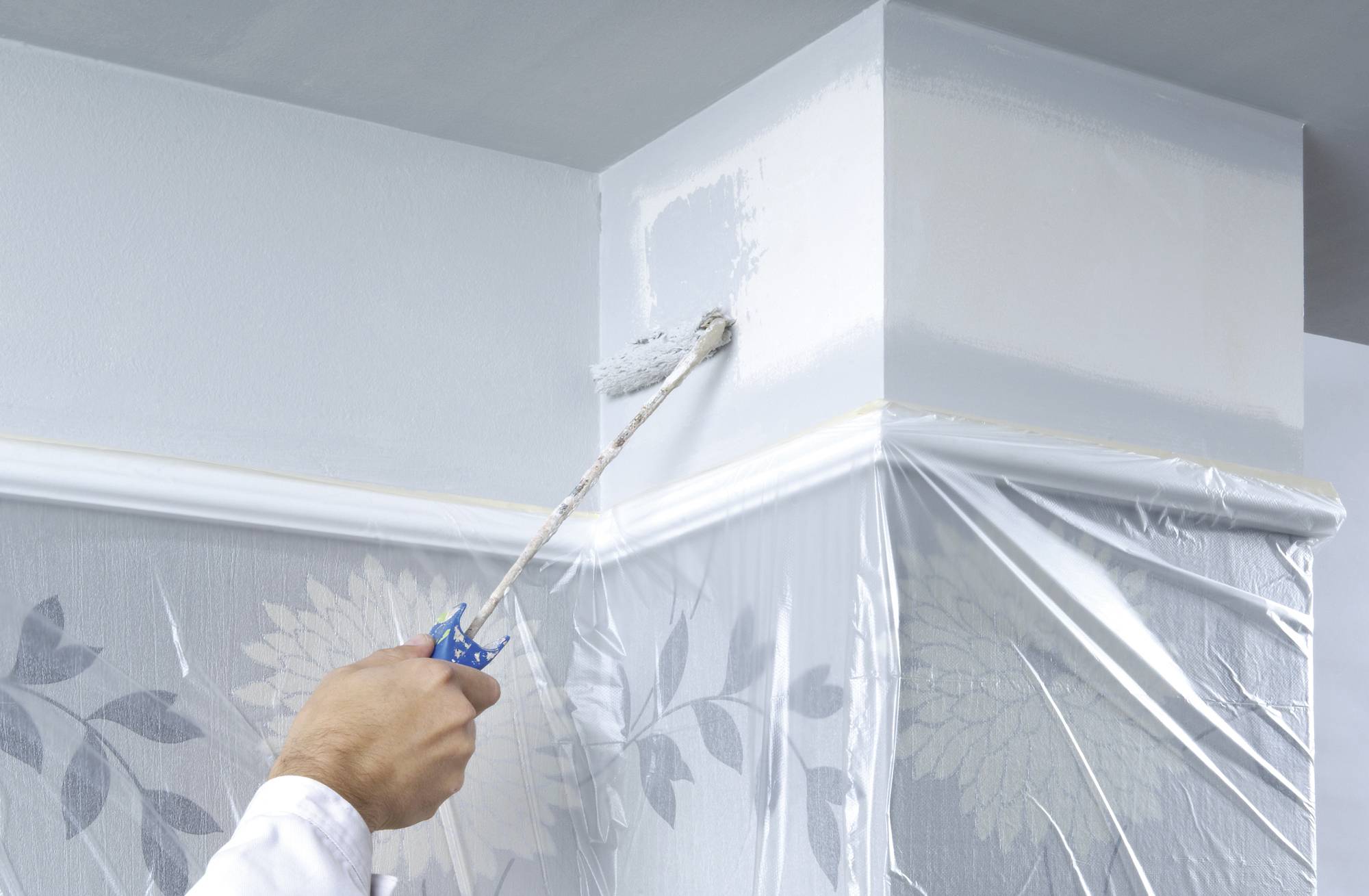Ein Handwerker streicht eine hellblaue Wand mit einer Rolle. Die Wand ist zum Schutz teilweise mit Plastikfolie abgedeckt.