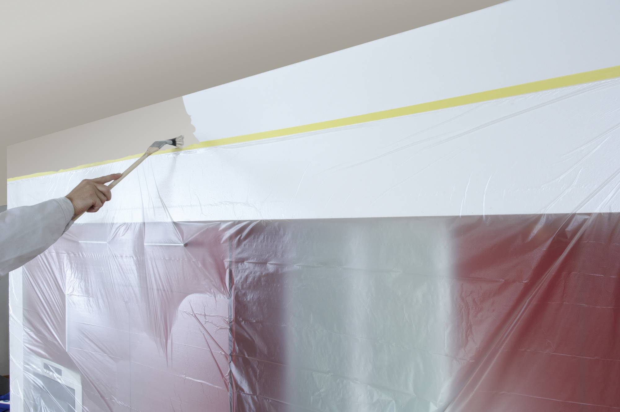 Eine Person streicht die Wand mit einem Pinsel weiß. Schutzfolie bedeckt die rote Wand darunter.