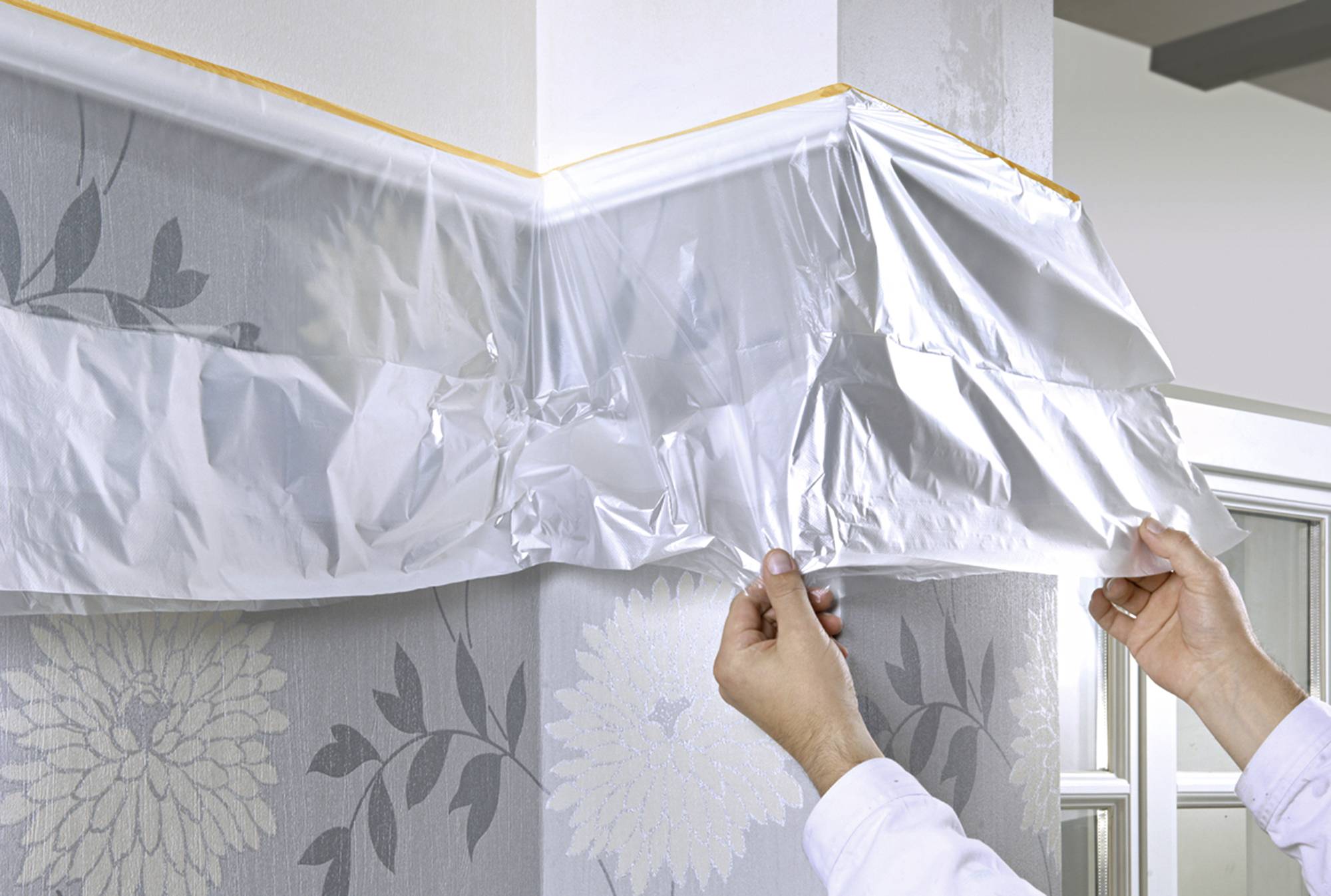 Eine Person bringt Schutzfolie an einer tapezierten Wand an, um vor Farbspritzern zu schützen. Die Wand hat ein florales Muster.