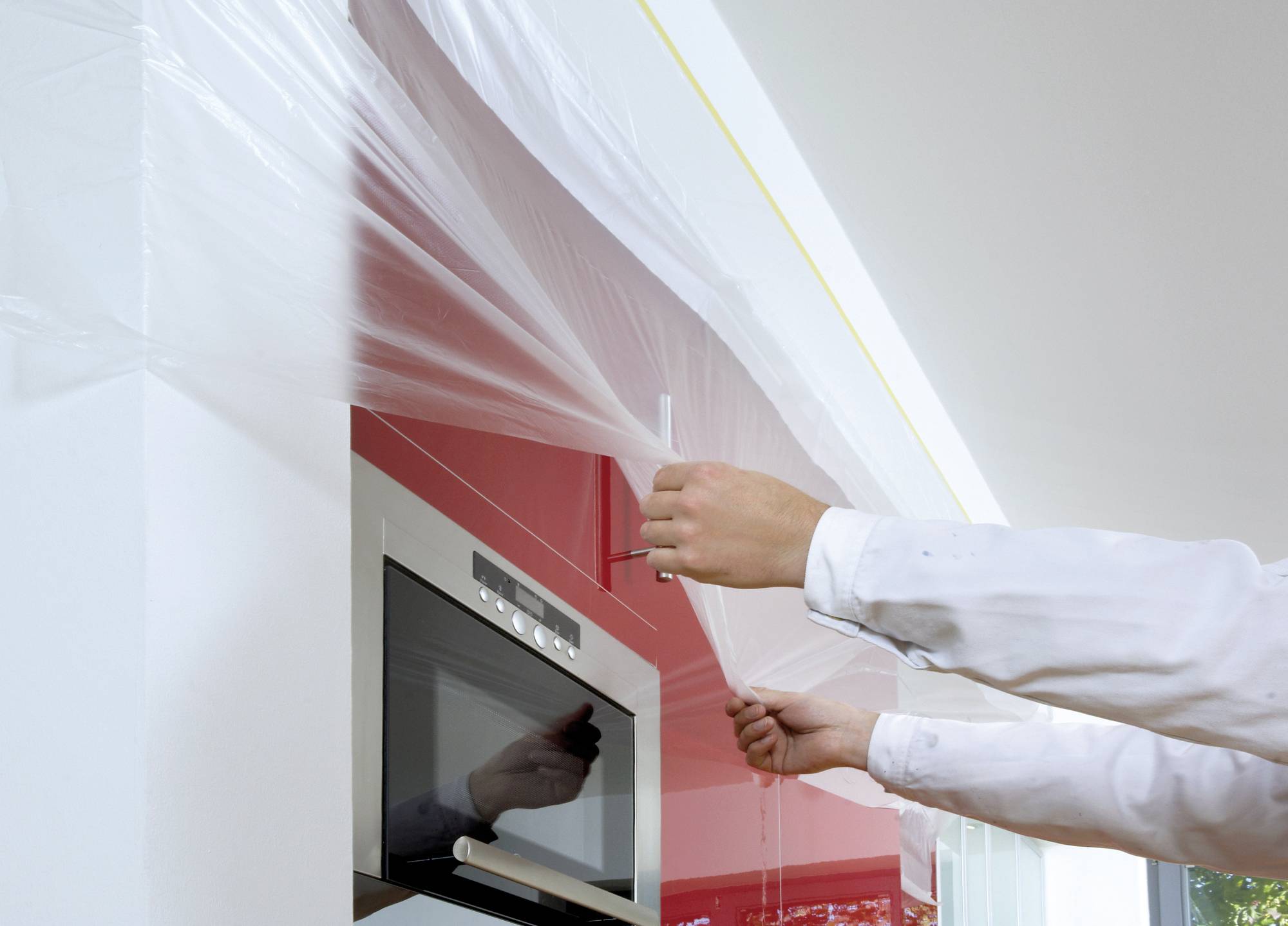 Eine Person in weißem Mantel zieht Plastikfolie über eine rote Küchenwand, nahe eines eingebauten Ofens. Renovierungsarbeiten finden statt.