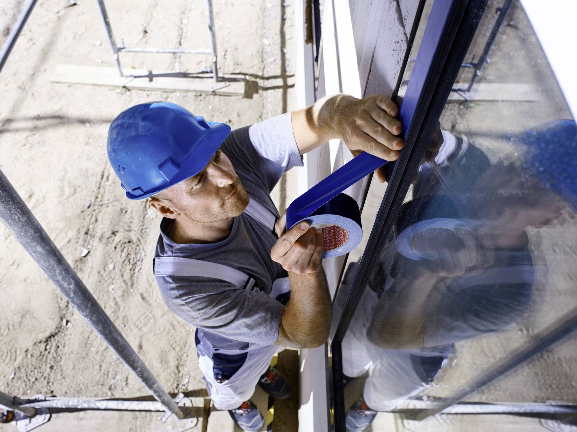 Ein Bauarbeiter mit blauem Schutzhelm klebt mit Klebeband ein Fenster ab. Er steht auf einem Gerüst und führt präzise Arbeiten aus.