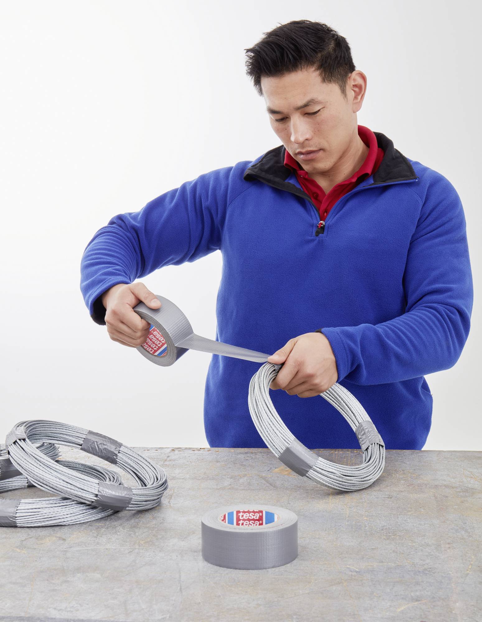 Eine Person wickelt graues Klebeband um einen Draht. Mehrere Drahtbündel und eine Rolle Klebeband liegen auf einem Tisch.