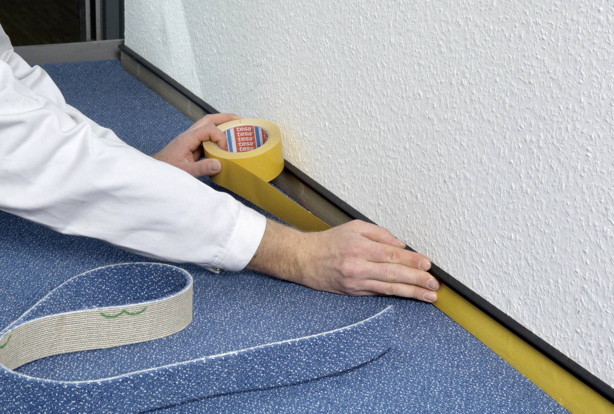 Eine Person klebt an einer Wand in der Nähe eines Teppichbodens gelbes Klebeband an, um einen sauberen Anstrich oder Schutz zu gewährleisten.