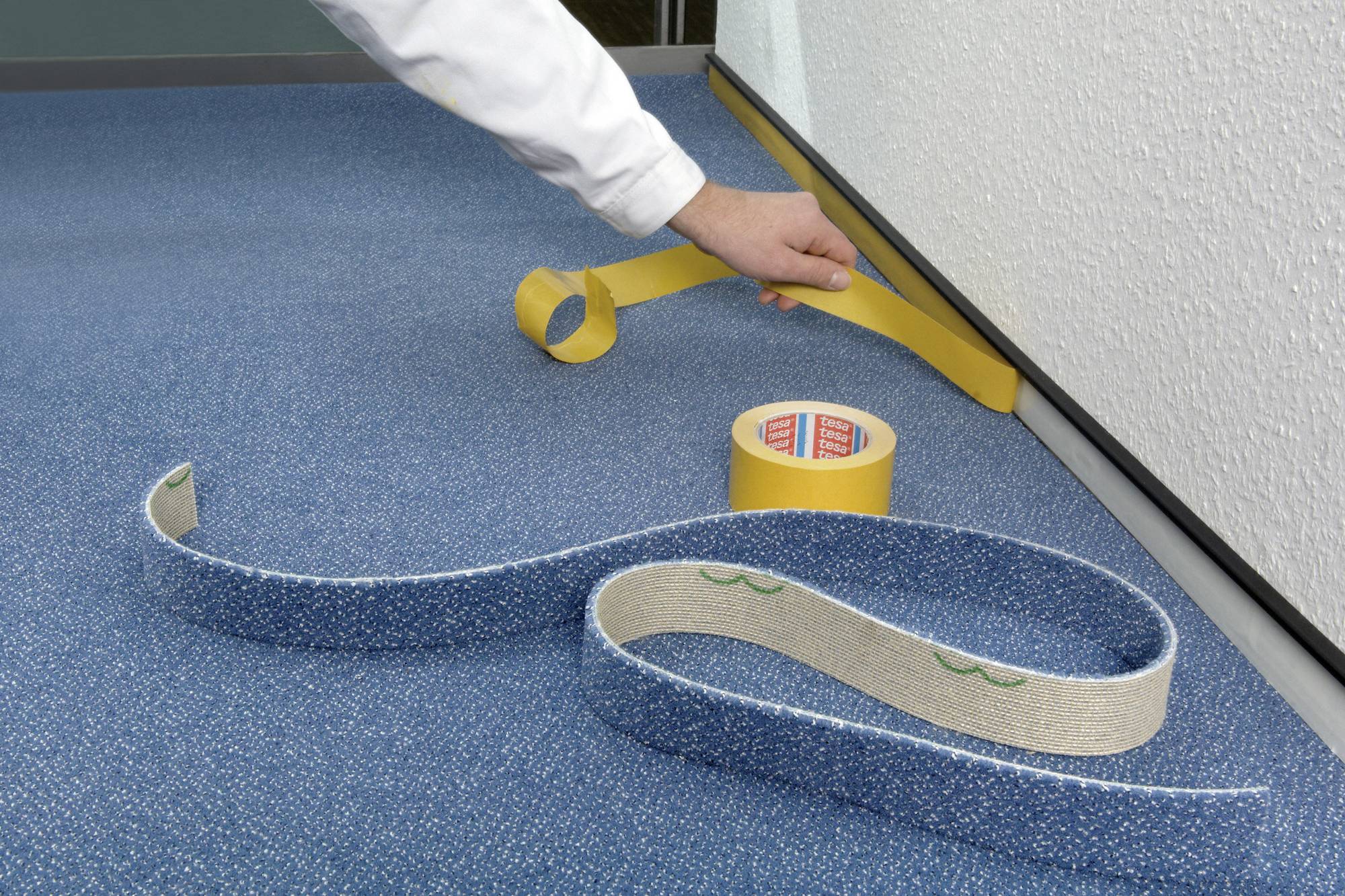 Person verlegt gelben doppelseitigen Klebeband-Streifen auf blauem Teppichboden, um diesen an einer weißen Wand zu befestigen.