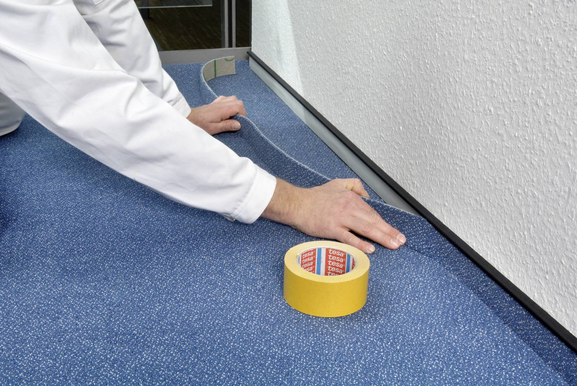 Eine Person verlegt blauen Teppichboden entlang einer Wand. Auf dem Boden liegt eine Rolle doppelseitiges Klebeband.