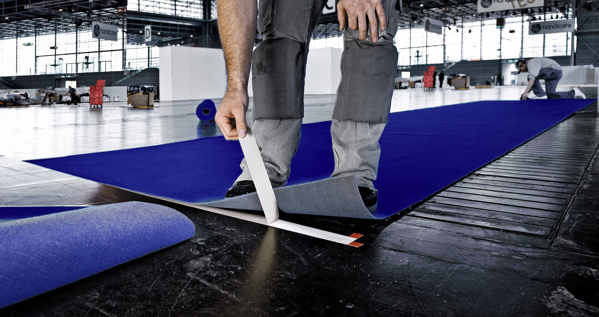 Person verlegt blauen Teppichboden in großer Halle. Ein Mitarbeiter arbeitet im Hintergrund.
